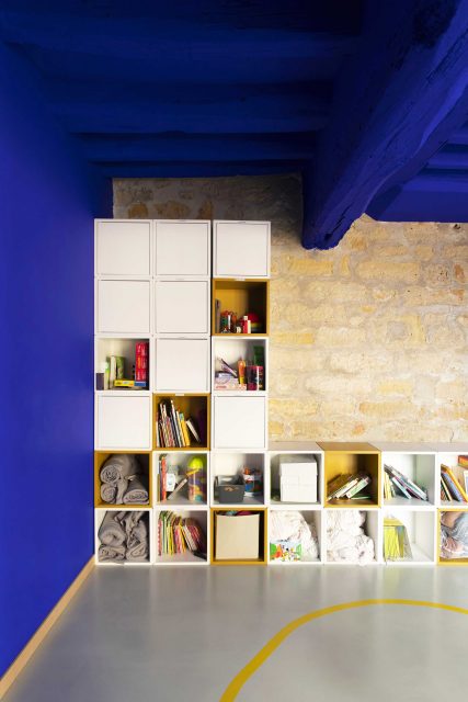 Vue de l'espace dortoir du projet de l'Ecole Montessori Square Paris 18 avec un focus sur le mur en pierres apparentes associé au fameux Bleu Klein très puissant de Ressource Peinture sur le plafond et dynamisé par l'ajout de rangements jaune ocre.