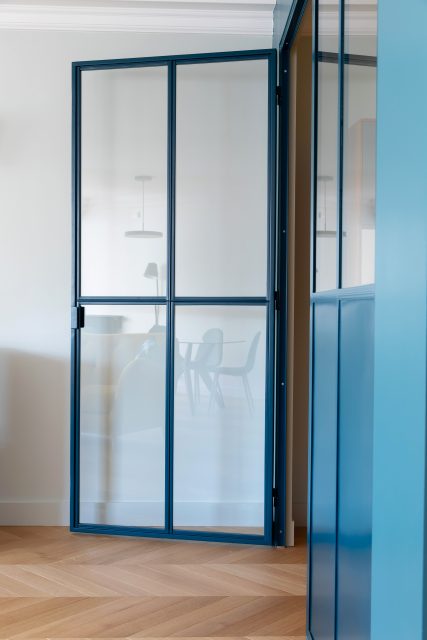 Détail de la porte vitrée de la verrière sur mesure bleue délimitant le bureau et son reflet de l'espace de vie attenant du projet Paris 16.