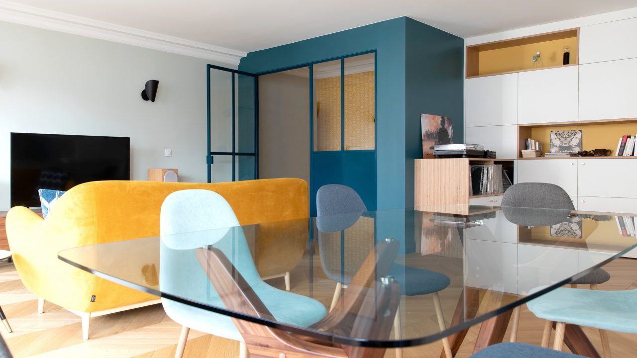 Vue de l'ensemble de l'espace salon / salle à manger du projet Paris 16 avec l'alcôve bureau vitrée bleue et son papier peint jaune en résonance avec le canapé jaune.