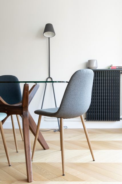 Vue de l'espace salle à manger minimaliste et contrastée avec sa table verre et noyer AMPM, ses chaises en tissu grises ainsi que son radiateur anthracite se détachant sur le mur blanc, Projet Paris 16