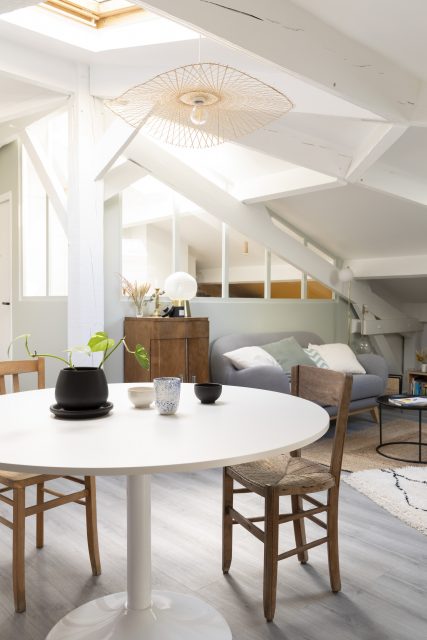 Vue sur l'espace salle à manger et sa jolie table ronde réchauffée de chaises chinées en bois avec en arrière plan l'alcôve bureau et sa verrière en bois peinte se nichant contre la structure insolite des combles du projet Ribera Paris 16.