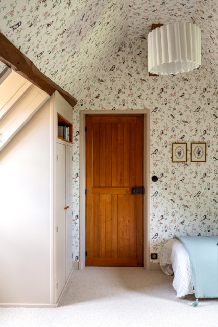 Une chambre lumineuse et chaleureuse avec des murs ornés de papier peint à motifs floraux et oiseaux sur fond vert pâle, rappelant les dortoirs d'antan et les maisons de notre enfance. Une porte en bois massif au style rustique, encadrée par des poutres apparentes, ajoute un supplément d'âme à cet espace empreint de nostalgie. Un lit simple, paré d'une couverture vert menthe et de draps vichy, évoque une douceur intemporelle.