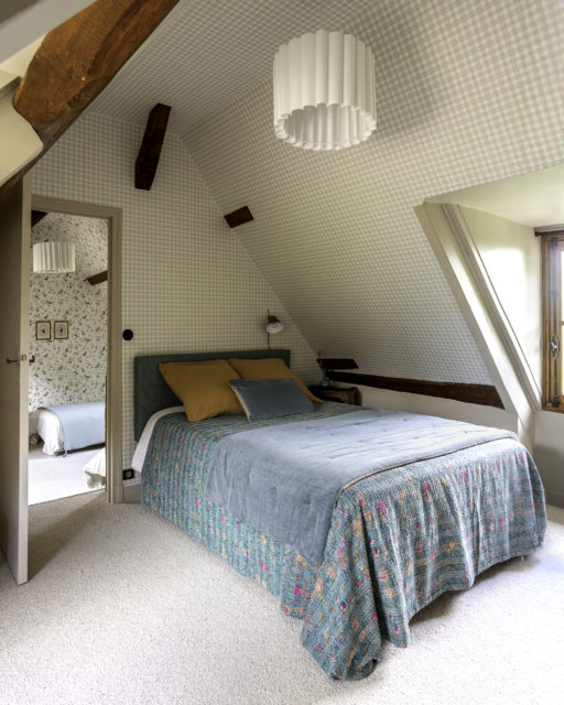 Une chambre mansardée lumineuse, décorée d'un papier peint vichy beige et blanc, avec un lit double habillé d'un couvre-lit bleu-vert à motifs fleuris. La tête de lit rembourrée dans une teinte vert sauge et les coussins jaunes et bleu pastel apportent une harmonie de couleurs douces. Une suspension plissée blanche et des poutres apparentes accentuent le charme authentique de la pièce.