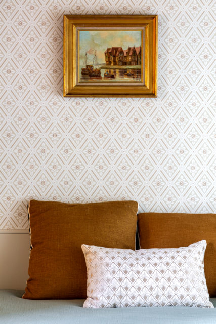 Détail d'une décoration intérieure dans une maison normande rénovée en bord d'eau par Ingrid Marcille. Un tableau encadré au style classique est suspendu au-dessus d'un mur tapissé de motifs géométriques rétro, accompagné de coussins dans des tons bruns et motifs assortis, créant une ambiance chaleureuse et vintage.